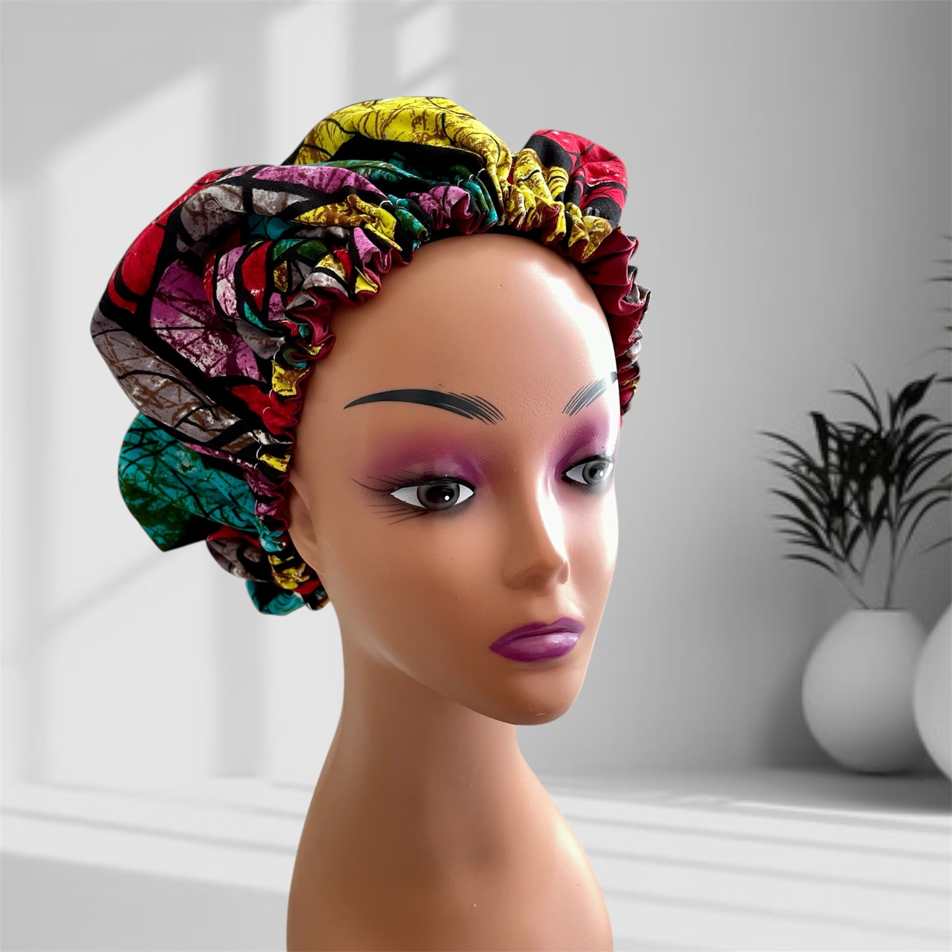 A mannequin head with bold makeup is shown wearing a colourful satin bonnet in shades of red, magenta and yellow with a leaf-pattern design, displayed indoors against a softly lit background with plants and vases.