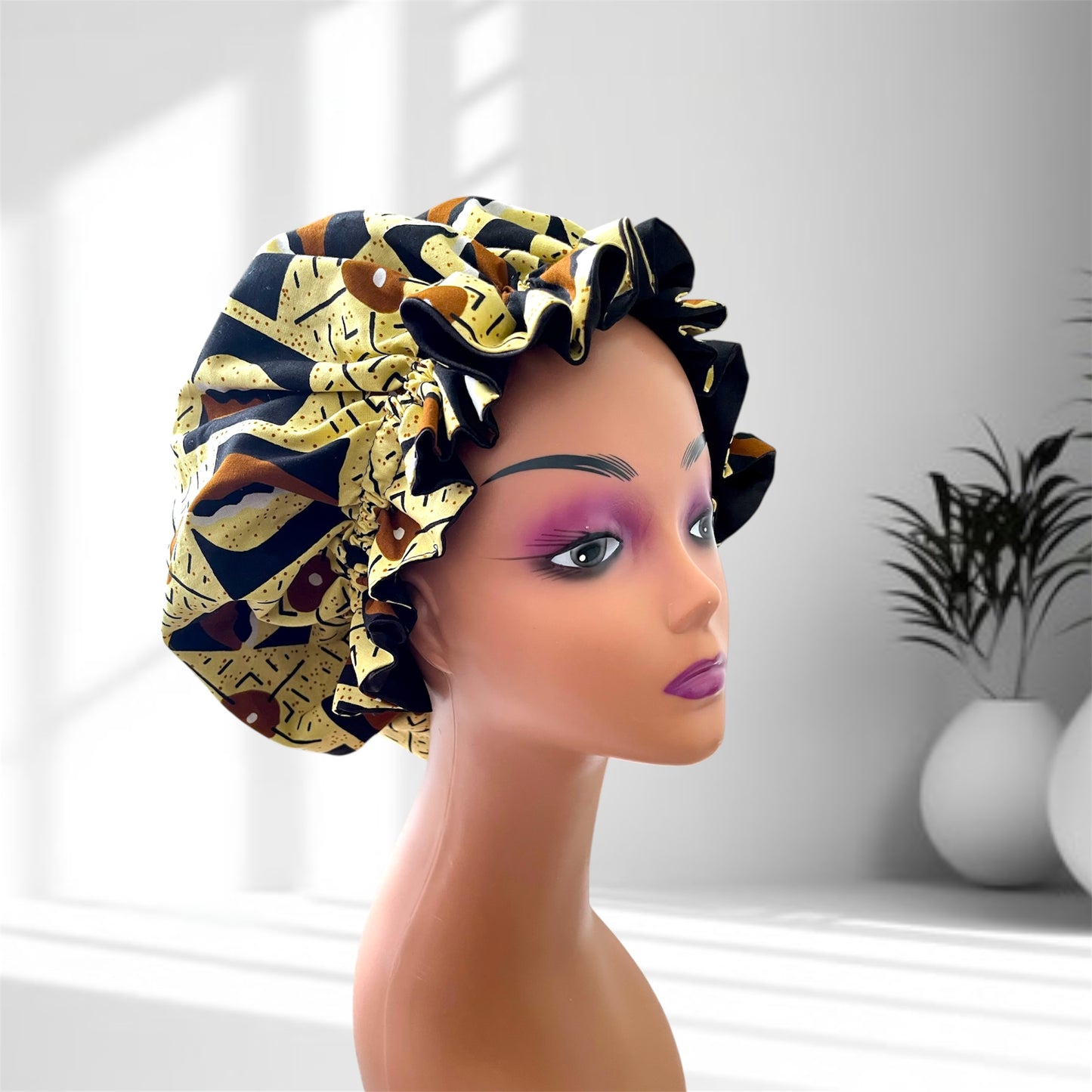 Stylish hair bonnet with a black, yellow, and brown geometric pattern displayed on a mannequin head, set against a softly lit indoor background with potted plants.