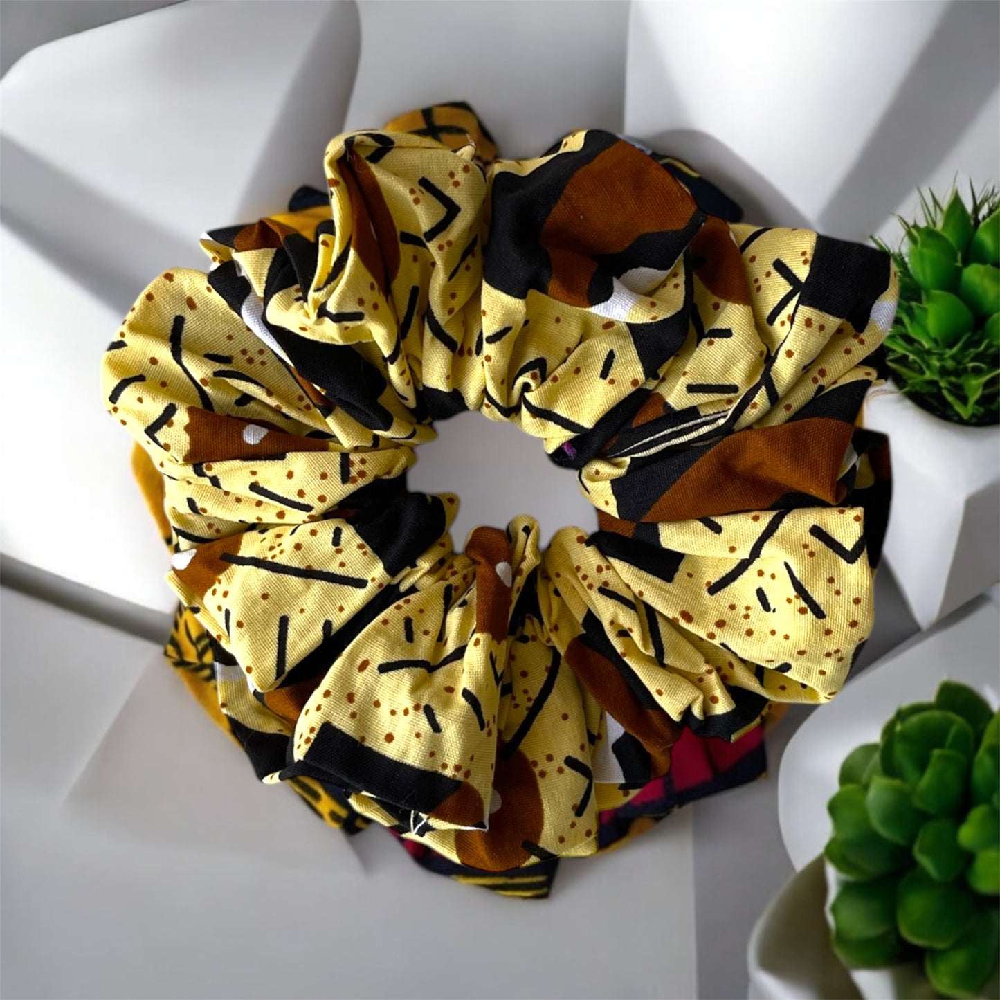 Scrunchies for the hair made with a yellow fabric featuring black, brown, and white abstract patterns, displayed on a white surface near green plants.