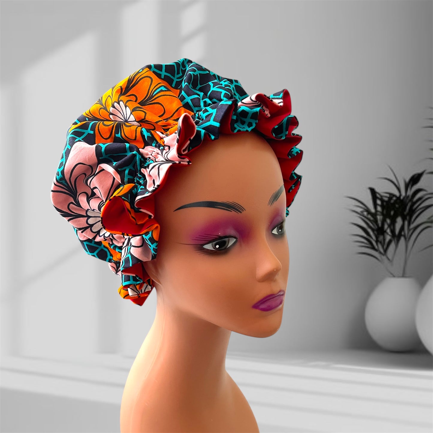 A mannequin head wearing a stylish hair bonnet with a colorful floral pattern, featuring shades of orange, pink, red, teal, yellow, and black, set against a blurred indoor background with potted plants.