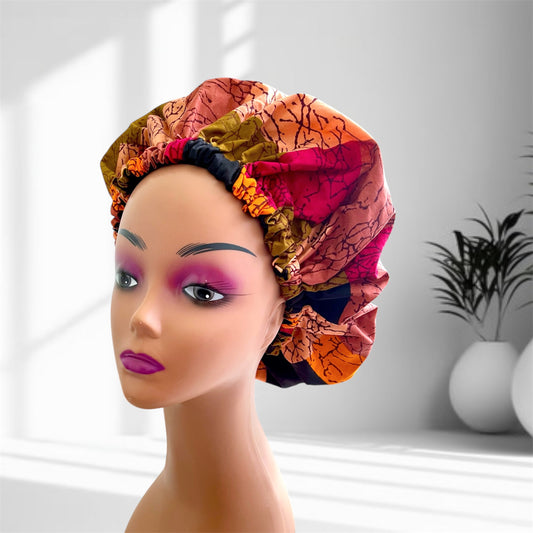 A mannequin head with bold makeup is shown wearing a colourful satin bonnet in shades of orange, pink, and brown with a leaf-pattern design, displayed indoors against a softly lit background with plants and vases.