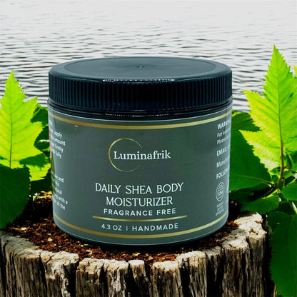 Luminafrik Daily Shea Body Moisturizer jar on a wooden block with leaves in the background