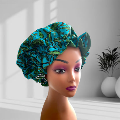 A mannequin head wearing a stylish hair bonnet in teal with green and black patterned designs, displayed against a blurred indoor background with potted plants.