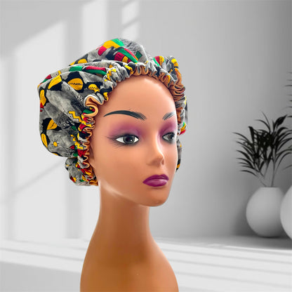 A mannequin head with bold makeup is shown wearing a colourful satin bonnet in shades of green, yellow, grey amd red with a leaf-pattern design, displayed indoors against a softly lit background with plants and vases.