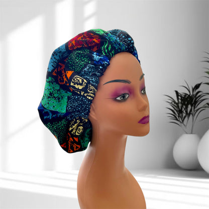 A mannequin head with bold makeup is shown wearing a colourful satin bonnet in shades of blue, green, orange and yellow with a leaf-pattern design, displayed indoors against a softly lit background with plants and vases.