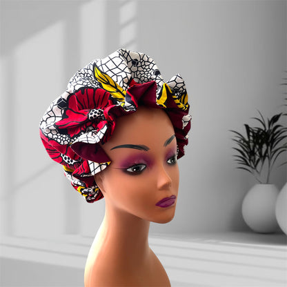 A mannequin head wearing a stylish hair bonnet with a vibrant design featuring red, yellow, black, and white colors, displayed against a blurred indoor background with potted plants.