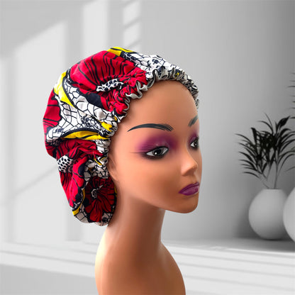 A mannequin head with bold makeup is shown wearing a colourful satin bonnet in shades of white, red and yellow with a leaf-pattern design, displayed indoors against a softly lit background with plants and vases.