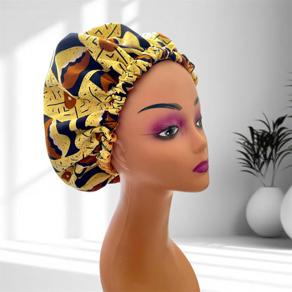 A mannequin head with bold makeup is shown wearing a colourful satin bonnet in shades of yellow, brown and blue with a leaf-pattern design, displayed indoors against a softly lit background with plants and vases.