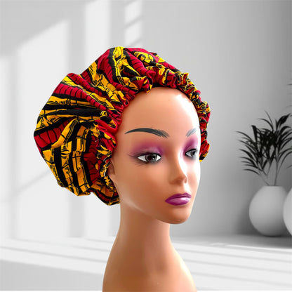 A mannequin head with bold makeup is shown wearing a colourful satin bonnet in shades of red, yellow and black with a leaf-pattern design, displayed indoors against a softly lit background with plants and vases.