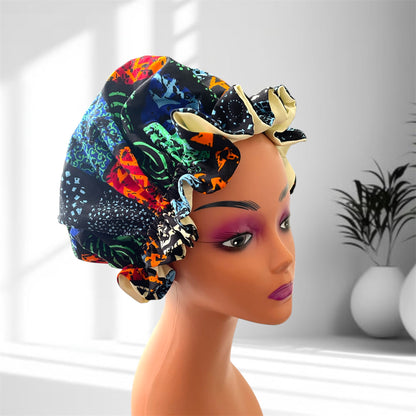 A mannequin head wearing a stylish hair bonnet with a vibrant multicolor patchwork design in shades of blue, green, red, orange, black, and cream, set against a blurred indoor background with potted plants.