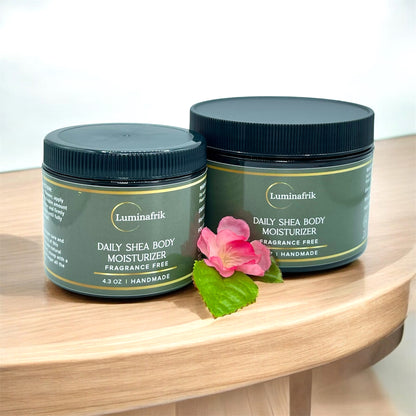 Two jars of Luminafrik Daily Shea Body Moisturizer on a wooden surface with a pink flower.
