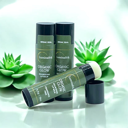 Several tubes of Luminafrik Organic Glow nourishing lip balm with green labels and black caps are displayed on a glossy surface, with one tube open to reveal the balm, set against a background decorated with green succulents.