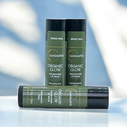 Three tubes of Luminafrik Organic Glow nourishing lip balm with green labels and black caps are displayed on a glossy surface, two standing upright and one lying horizontally in front, with a softly blurred blue and white background.