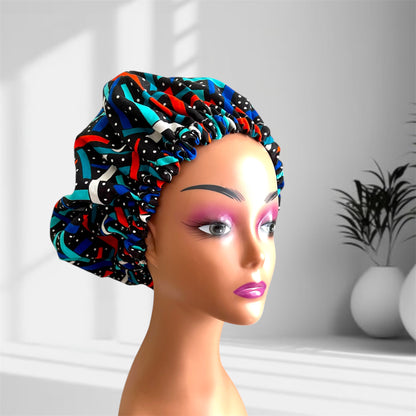 A mannequin head with bold makeup is shown wearing a colourful satin bonnet in shades of black, blue, whit with a leaf-pattern design, displayed indoors against a softly lit background with plants and vases. and red